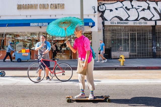 Older Asian woman wearing tan pants and a hot pink shirt, holding a turquoise paper umbrella while skateboarding down the street.